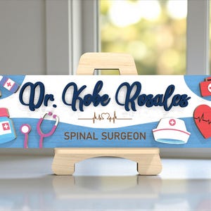 May include: A wooden sign on a small easel, featuring the name "Dr. Kobe Rosales" in navy blue script. The sign includes medical-themed icons like a stethoscope, heart, and medical kit, with the words "Spinal Surgeon" below.