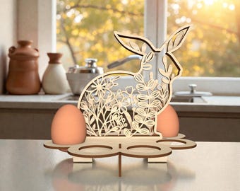 May include: Wooden Easter egg holder in the shape of a rabbit with floral cutouts. The holder has spaces for two eggs and is decorated with two orange eggs. The design is made of light-colored wood.