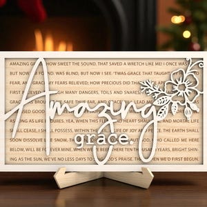 Puede incluir: Un letrero de madera con las palabras "Amazing Grace" recortadas en escritura blanca. El letrero tiene un diseño floral y un fondo de texto impreso. Se muestra sobre un soporte de madera.