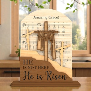 May include: Wooden religious ornament with a cross design, the text "Amazing Grace!" and "He is not here, He is Risen." The layered design incorporates musical notes and a landscape, with a natural wood finish.
