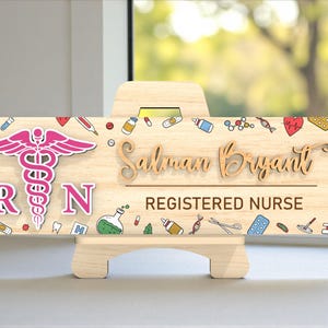 May include: A wooden Registered Nurse sign with a pink caduceus symbol and the letters "RN". The name "Salman Bryant" is displayed in a cursive font. The sign is decorated with medical-themed illustrations and sits on a wooden easel.