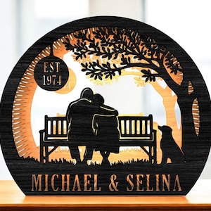 Pode incluir: Peça decorativa de madeira preta e castanha com um casal num banco com um cão debaixo de uma árvore. O texto "EST 1974" está num círculo, e os nomes "MICHAEL & SELINA" estão na parte inferior. A peça é cortada a laser.