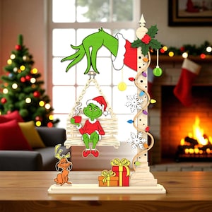 May include: Wooden Christmas decoration featuring a green character in a Santa hat, holding a mug, seated on a swing. The decoration includes a reindeer, presents, and a string of lights. The background shows a Christmas tree and a fireplace.
