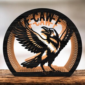 May include: A layered wooden sculpture of a raven with outstretched wings, set within a circular frame. The raven is black and orange, with the word "CAW" visible at the top. The sculpture is on a wooden surface.