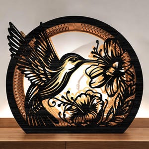 Könnte beinhalten: Eine schwarze und braune Holz-Hummingbird-Lampe. Die Lampe hat ein detailliertes Ausschnittdesign eines Kolibris und Blumen in einem kreisförmigen Rahmen. Die Lampe steht auf einer Holzoberfläche.