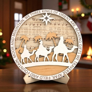 May include: A wooden Christmas decoration with a circular design. It features the text "We Three Kings of Orient Are," silhouettes of three kings on camels, palm trees, and musical notes. The decoration is mounted on a wooden stand.
