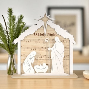 May include: A wooden nativity scene with a star on top, featuring cut-out figures of Mary, Joseph, and baby Jesus. The background is sheet music with the words "O Holy Night". The scene is set on a wooden base.