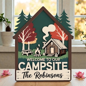 May include: A layered wooden sign shaped like a house, depicting a campsite with a cabin, trees, a picnic table, and a campfire. The sign displays the text "WELCOME TO OUR CAMPSITE" and "The Robinsons."