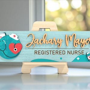 May include: A wooden desk sign for a Registered Nurse, featuring the name "Zachary Mayer" in a cursive font. The sign has a teal background with medical-themed graphics, including a stethoscope, heart, and nurse's cap.