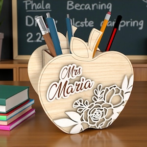 Puede incluir: Organizador de escritorio de madera con forma de manzana con el nombre "Mrs. Maria" en cursiva. El organizador tiene diseños florales recortados y contiene bolígrafos y lápices. Una pila de libros y una planta en maceta están en el fondo.