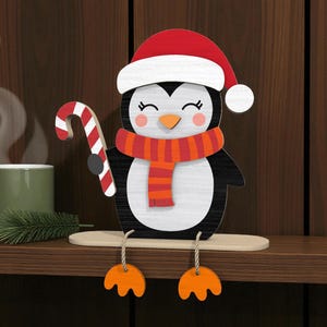 May include: A wooden penguin Christmas decoration wearing a red and white Santa hat and an orange and red scarf. The penguin holds a candy cane and has orange feet. A green mug with steam and a sprig of pine are in the background.