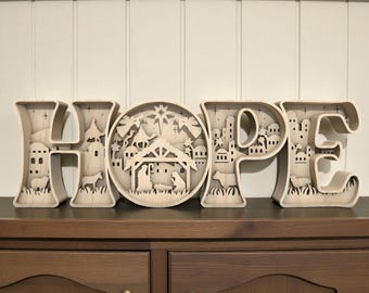 May include: Wooden decorative sign spelling "HOPE" with a nativity scene in the "O". The letters are light brown and feature cut-out designs of buildings, animals, and stars. The sign is displayed on a dark brown wooden surface.