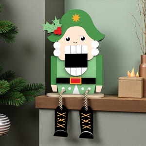 May include: A wooden Christmas nutcracker figurine in green, white, and black. The figurine has a green hat with a gold star and holly leaves. Black boots hang from the figurine. A Christmas tree and ornaments are in the background.