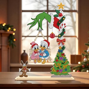 May include: A wooden Christmas decoration featuring cartoon characters. Donald and Daisy Duck are seated on a swing, while a reindeer stands below. A Christmas tree and a Grinch hand complete the festive scene. The decoration is set on a wooden surface.