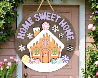 Könnte beinhalten: Runder Holzschild mit dem Schriftzug "HOME SWEET HOME" in Lavendelfarben. Es zeigt ein Lebkuchenhaus-Design mit Süßigkeiten und Schneeflocken. Ein Seil ist zum Aufhängen befestigt.