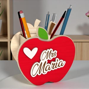 Puede incluir: Un organizador de escritorio en forma de manzana roja con un corazón blanco y el nombre "Mrs. Maria" en cursiva. El organizador de madera contiene bolígrafos y lápices. Una hoja verde está unida a la parte superior de la manzana.