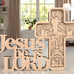 May include: A wooden cross with layered cutouts depicting religious scenes, including Jesus and the crucifixion. The words "Jesus he is LORD" are cut out in a stylized font. The cross is made of light-colored wood and stands on a flat surface.