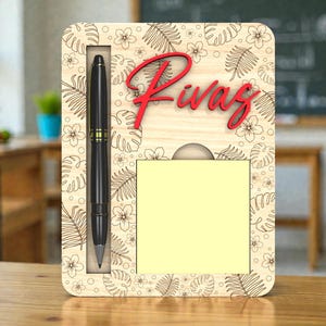 Op de afbeelding: Houten bureau-organizer met een pen, notitieblok en de naam "Rivas" in het rood. De organizer heeft een bloemen- en bladerenpatroon. Een zwarte pen met gouden accenten zit in een sleuf. Een geel notitieblok staat eronder.