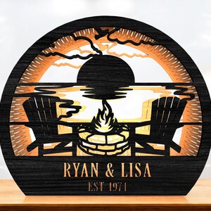 Könnte beinhalten: Ein schwarzes Holzdekorstück mit einer Sonnenuntergangsszene, zwei Stühlen und einem Lagerfeuer. Die Namen "RYAN & LISA" und das Jahr "EST 1971" sind eingraviert. Das Design ist mit orangefarbenen und gelben Akzenten versehen.