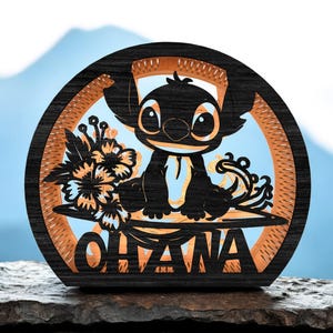 Peut inclure: Une œuvre d'art en bois ronde et superposée représentant une silhouette noire d'un personnage de dessin animé aux grands yeux, assis avec des fleurs. Le mot "OHANA" est découpé en bas. Le fond est bleu clair avec une montagne au loin.