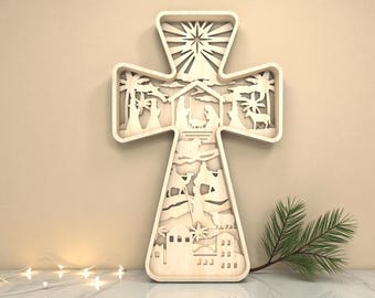 May include: A wooden cross-shaped wall hanging with a layered design depicting a nativity scene. The cross features cut-out silhouettes of palm trees, figures, and buildings. The natural wood color contrasts with the detailed interior scenes.