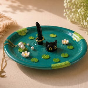 May include: A round teal ceramic jewellery dish featuring a black cat head and a black cat tail rising from the centre. The surface is decorated with small green lily pads and white lotus flowers. A gold-toned necklace with a crescent moon pendant rests on the dish, and a gold ring is placed around the cat tail.