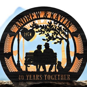 Può includere: Targa rotonda in legno con bordo nero e design intagliato. Il design presenta sagome di una coppia su una panchina, un albero e un lampione. Il testo include "ANDREW & KAYLIN", "1974" e "40 YEARS TOGETHER".