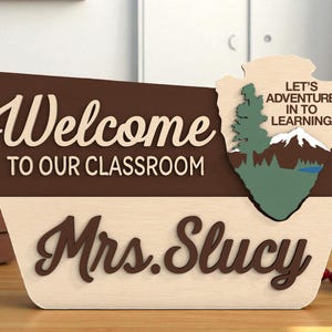 Puede incluir: Letrero de aula de madera en marrón y beige. El letrero dice "Bienvenido a nuestra clase" y "Sra. Slucy" en una fuente decorativa. Un gráfico de montaña y árbol está a la derecha, con el texto "Aventurémonos en el aprendizaje".