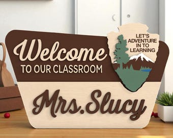 Puede incluir: Letrero de aula de madera en marrón y beige. El letrero dice "Bienvenido a nuestra clase" y "Sra. Slucy" en una fuente decorativa. Un gráfico de montaña y árbol está a la derecha, con el texto "Aventurémonos en el aprendizaje".