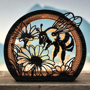 May include: A wooden, layered art piece featuring a bee hovering near sunflowers. The bee is black and yellow, with intricate wing details. The flowers are outlined in black, with a light-colored center. The frame is dark wood with a woven texture.