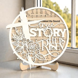 May include: A circular wooden sign featuring a cross, floral cutouts, and the words "true STORY HE IS RISEN". The sign is displayed on a wooden stand. The background includes sheet music.