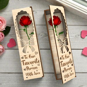 Puede incluir: Dos cajas de regalo de madera con una rosa roja en su interior. Las cajas tienen un diseño decorativo recortado con un lazo y las palabras "To Our Favorite Person With Love". Decoraciones rosas en forma de corazón están dispersas.