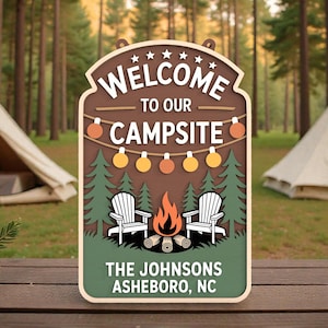 May include: A wooden sign with a camping theme. The sign reads "WELCOME TO OUR CAMPSITE" with a graphic of a campfire, two chairs, and trees. The bottom of the sign says "THE JOHNSONS ASHEBORO, NC".