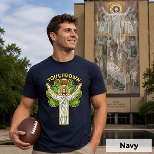 May include: Navy blue t-shirt with the word "Touchdown" above a graphic of a football and a religious figure in a stained-glass style design. The shirt is a solid navy color. A brown football is held in the person's hand.