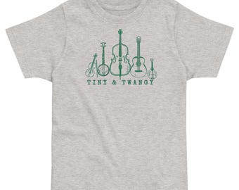 Könnte beinhalten: Hellgraues T-Shirt mit einer grünen Illustration von Streichinstrumenten, darunter Geige, Banjo, Cello, Gitarre und Mandoline. Der Text "TINY & TWANGY" ist darunter aufgedruckt.