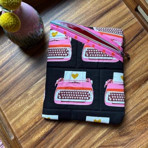 May include: A black quilted pouch with a pink zipper and a repeating pattern of pink typewriters with yellow hearts. The pouch is on a wooden tray with a pink vase and yellow flowers.