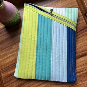 May include: A quilted laptop sleeve with a zipper closure. The sleeve features vertical stripes in shades of green, blue, and white. The zipper is silver, and the sleeve is on a wooden tray.