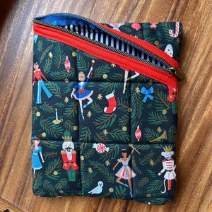 May include: A quilted, rectangular pouch with a zipper. The dark green fabric is patterned with Christmas-themed illustrations, including ballerinas, nutcrackers, and ornaments. The zipper is red with a black and white striped interior.