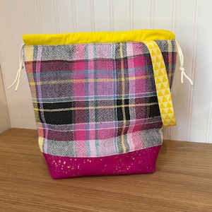 May include: A colorful tote bag with a plaid pattern in pink, purple, black, and yellow. The bag has a bright yellow top and a magenta base with gold flecks. A yellow strap with triangle designs is attached.