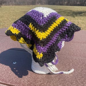 May include: A triangular crochet headscarf featuring a chevron pattern in alternating stripes of white, purple, black, and yellow yarn. The headscarf includes two long crocheted ties at the bottom corners for securing the accessory around the head.