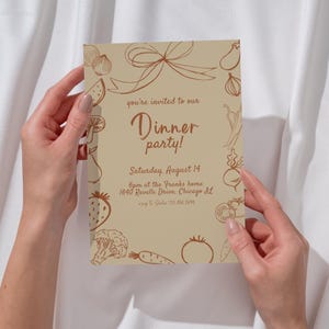 May include: A tan invitation card with a brown border of sketched vegetables and a bow. The text reads "Dinner party!" with the date, time, and location. The card is held by two hands against a white background.