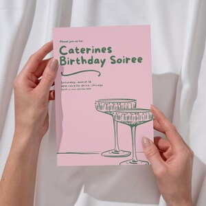 Könnte beinhalten: Eine rosa Einladung mit grünem Text und Illustrationen. Der Text lautet "Caterines Birthday Soiree" mit Veranstaltungsdetails. Zwei Champagnergläser sind unten abgebildet. Die Einladung wird von zwei Händen gehalten.