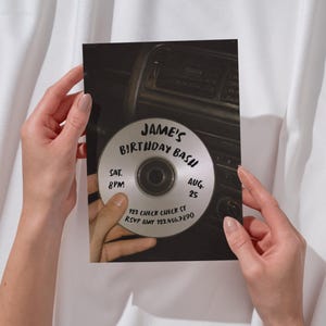 May include: A rectangular invitation card featuring a photographic print of a silver compact disc held against a car dashboard. The disc displays the text Jame's Birthday Bash, Sat. 8pm, Aug. 25, 123 Check Check St, and RSVP Amy 123.456.7890 in black ink.