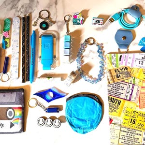 May include: A collection of items including a cassette tape pencil case, blue charging cable, beaded bracelet, concert ticket collage notebook, various keychains, pencils, nail files, a blue pouch, and small pins featuring music and smiley face themes.