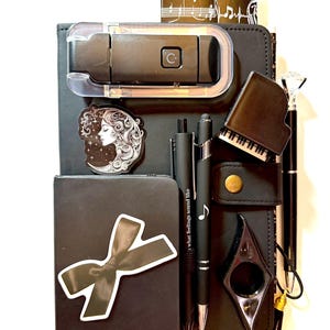 May include: A black notebook set featuring a black bow sticker, a celestial profile sticker, a black piano-shaped sharpener, two black pens with music notes, a black stapler, and a pen with a diamond-shaped topper. Music-themed paper accents are visible.