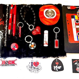 May include: A collection of music-themed items including a black Fender sock, red pens, a black beaded bracelet with a red heart charm, a red circular pouch, a black pouch with a cassette tape print, a USB cable, stickers, and keychains.