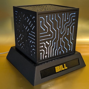 Puede incluir: Lámpara negra con forma de cubo con diseño de placa de circuito, iluminada con luz azul. La lámpara se apoya sobre una base negra con el nombre "BILL" en letras amarillas. Un artículo decorativo único.