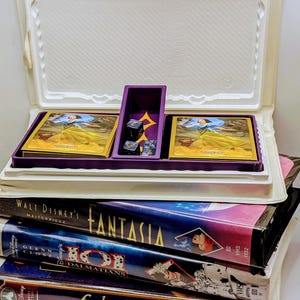 May include: A vintage board game with purple game pieces and cards, inside a white plastic case. Below the game are several VHS tapes, including "Fantasia", "101 Dalmatians", and "Lady and the Tramp".