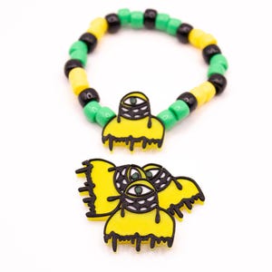 Puede incluir: Una pulsera de cuentas con cuentas verdes, amarillas y negras, combinada con un dije a juego. El dije es amarillo con un contorno negro y tres ojos.