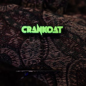 May include: A green and black pin with the word "CRANKOAT" in a bold, stylized font. The pin is set against a dark, patterned fabric background. The pin is likely a clothing accessory.